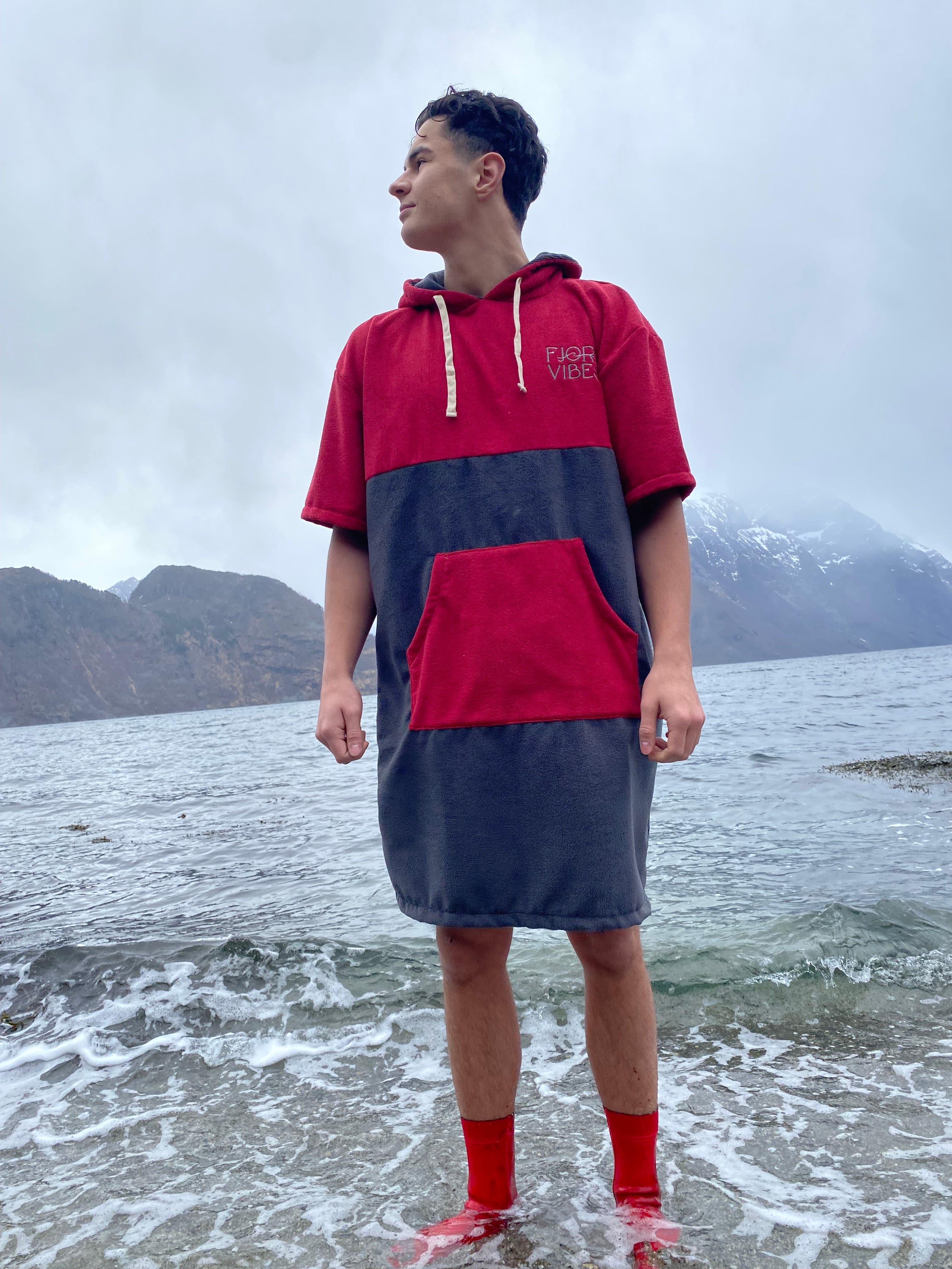 en mann som står i fjøra med vann til anklene og har på badeponcho i rødt og grått